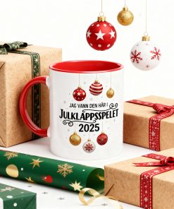 Vit kaffemugg med röd insida och handtag, tryckt med texten "Jag vann den här i Julklappsspelet 2025" och hängande julkulor.